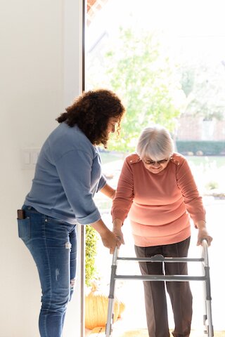 Pflegekraft unterstützt Seniorin mit Rollator beim Betreten des Hauses @ 